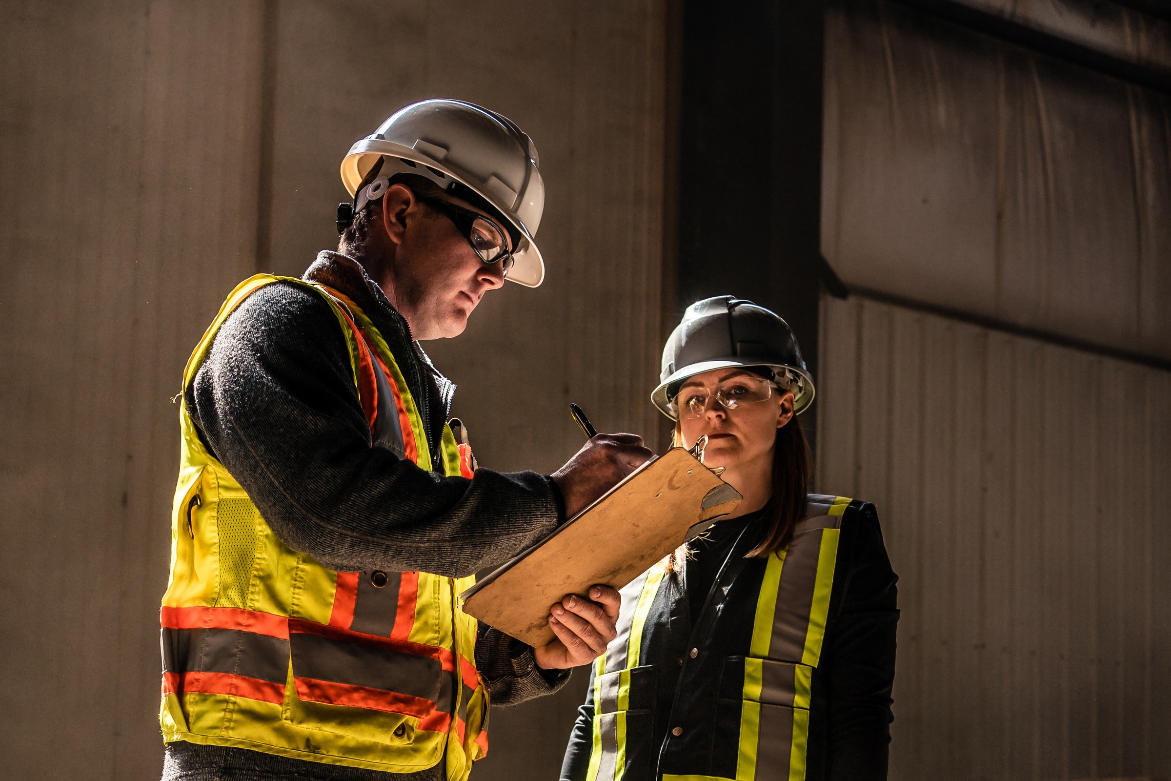 A COR auditor in a hard hat, safety glasses and hi-vis vest reviewing a clipboard with a client's safety lead in a sunlit shop.