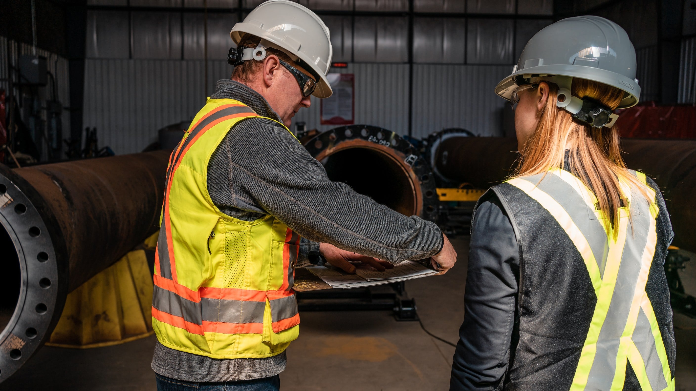 Two safety professionals in hard hats reviewing a document beside large pipe spools in a fabrication shop.