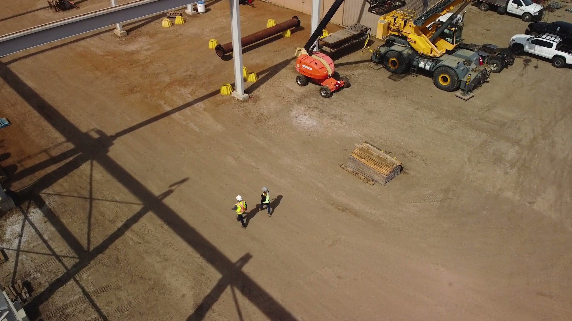 Aerial view of a fabrication yard with a boom lift, a crane and two workers in hi-vis walking between steel frames.