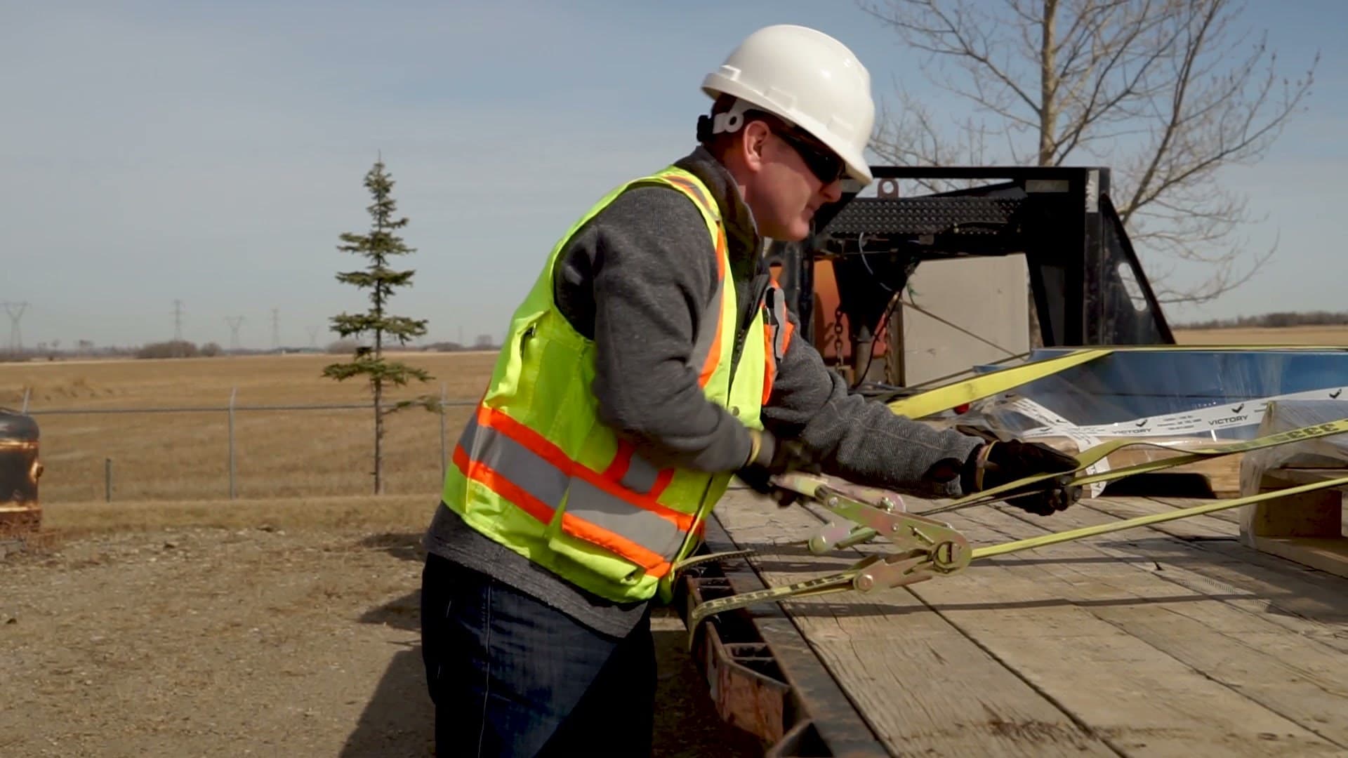 A worker in a hard hat, safety glasses and hi-vis vest tightening a cargo strap on a flatbed trailer load.