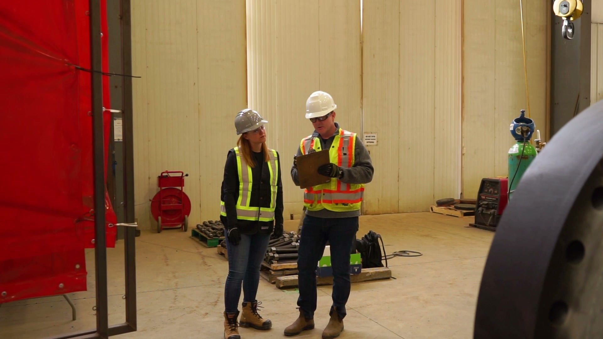 An auditor and a worker in hard hats and hi-vis vests talking beside a red welding curtain in a fabrication shop.