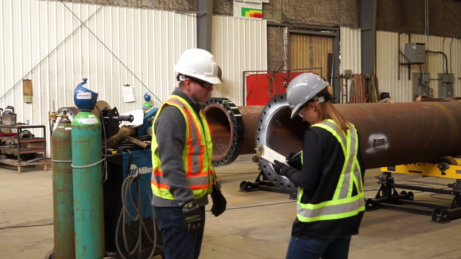 Two safety professionals in hard hats and hi-vis vests reviewing a checklist beside a pipe spool and gas cylinders in a fabrication shop.