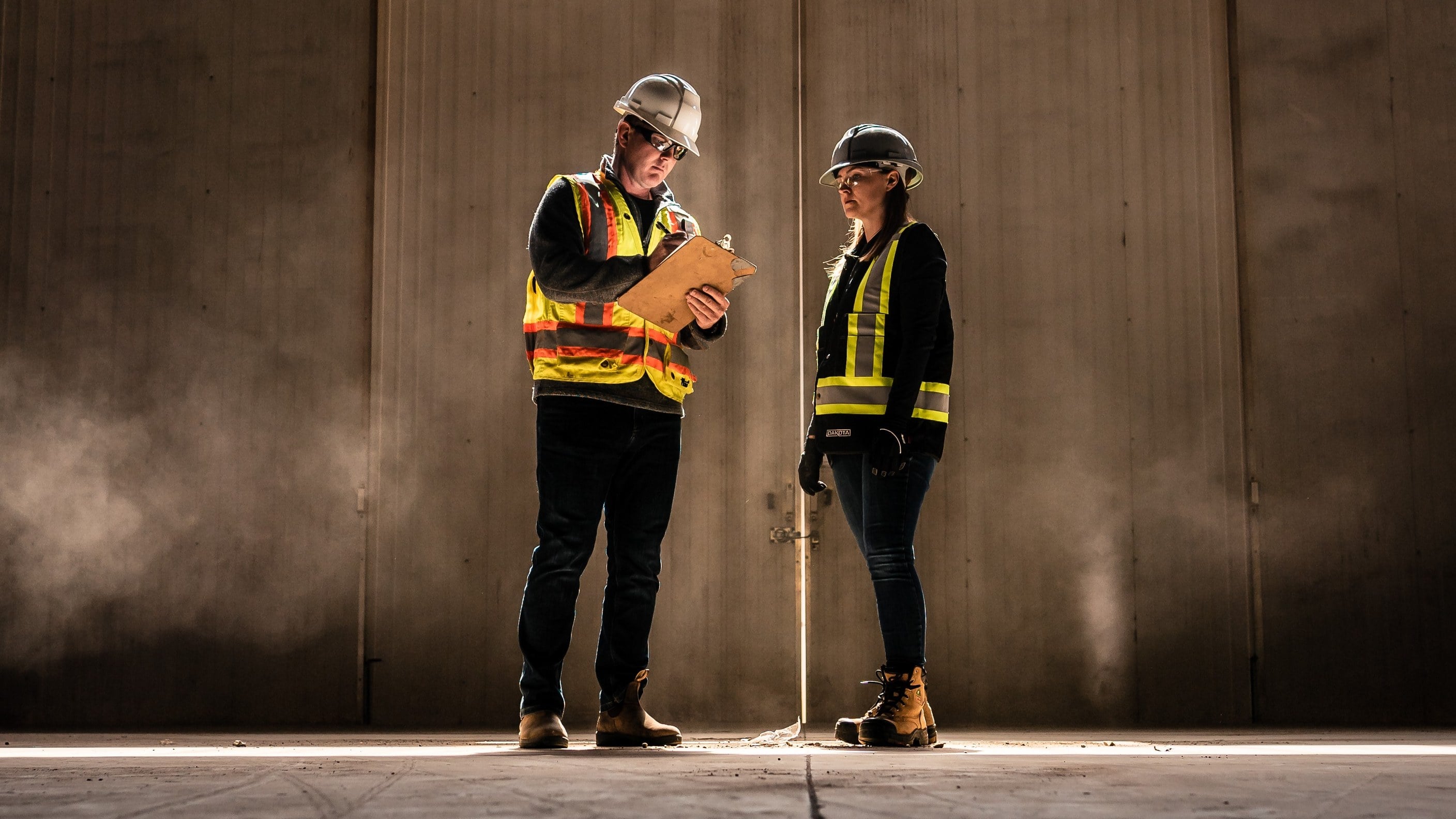 Two On-Track safety professionals in hard hats and hi-vis vests reviewing a clipboard in a sunlit industrial building.