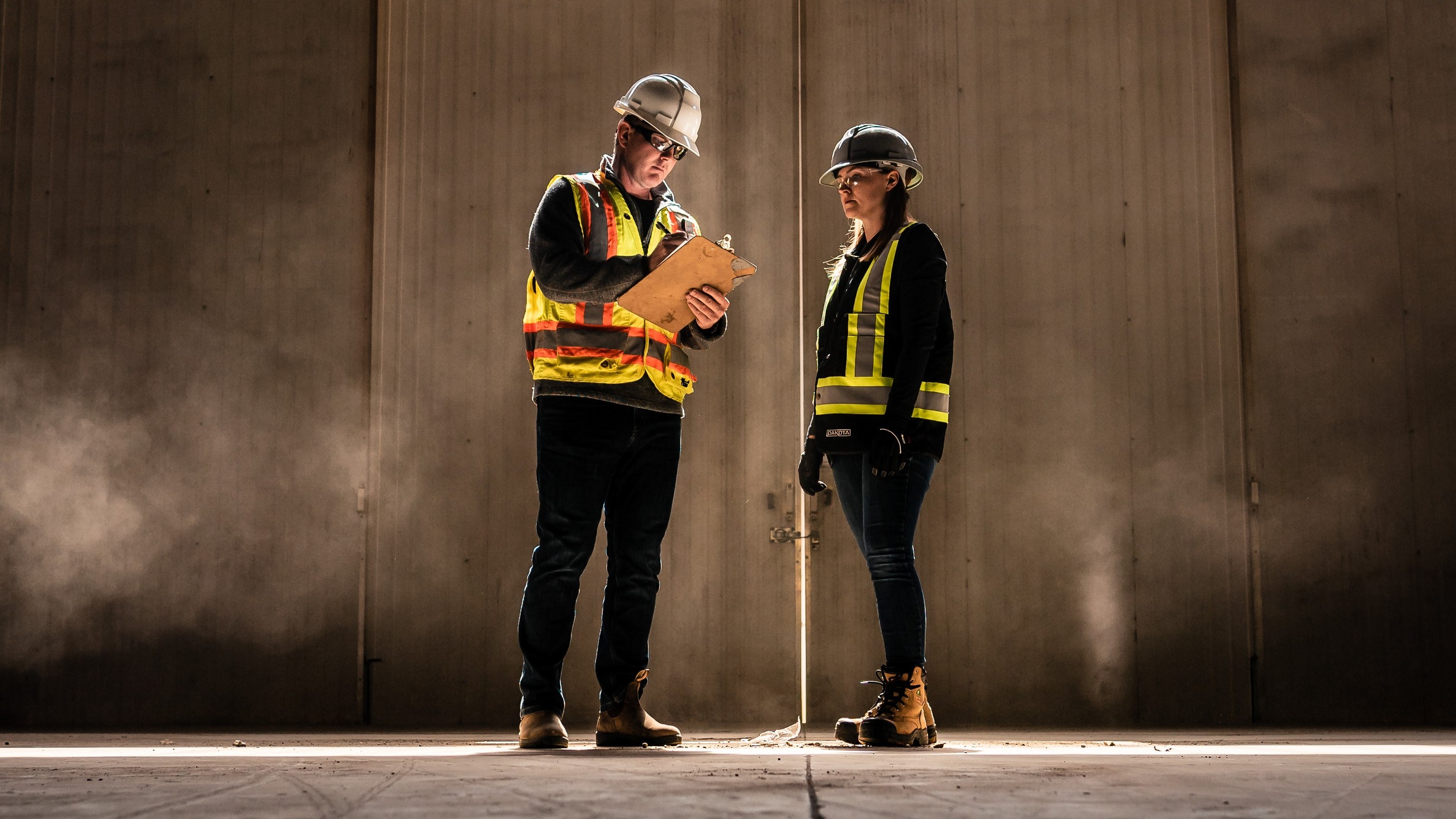 Two On-Track safety professionals in hard hats and hi-vis vests reviewing a clipboard in a sunlit industrial building.