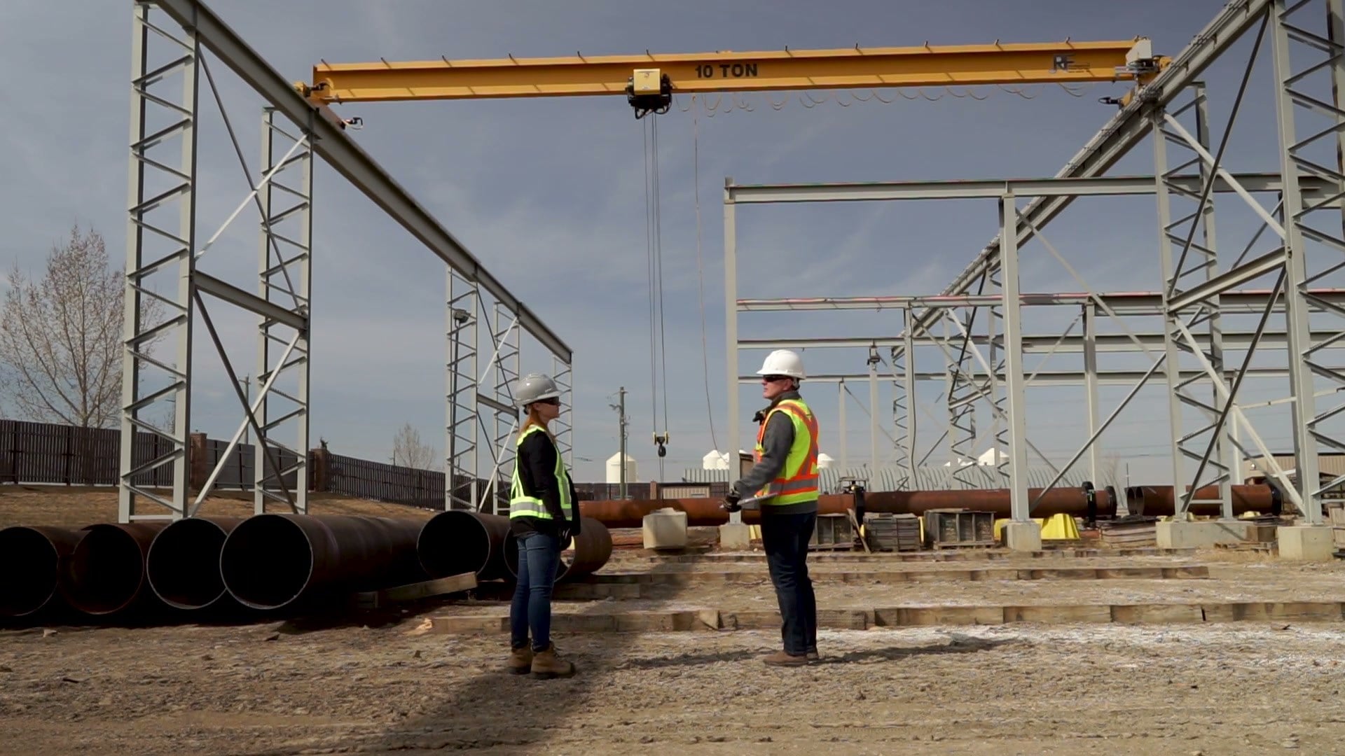 Two workers in hard hats and hi-vis vests in a pipe yard beneath a gantry crane, with steel frames behind.