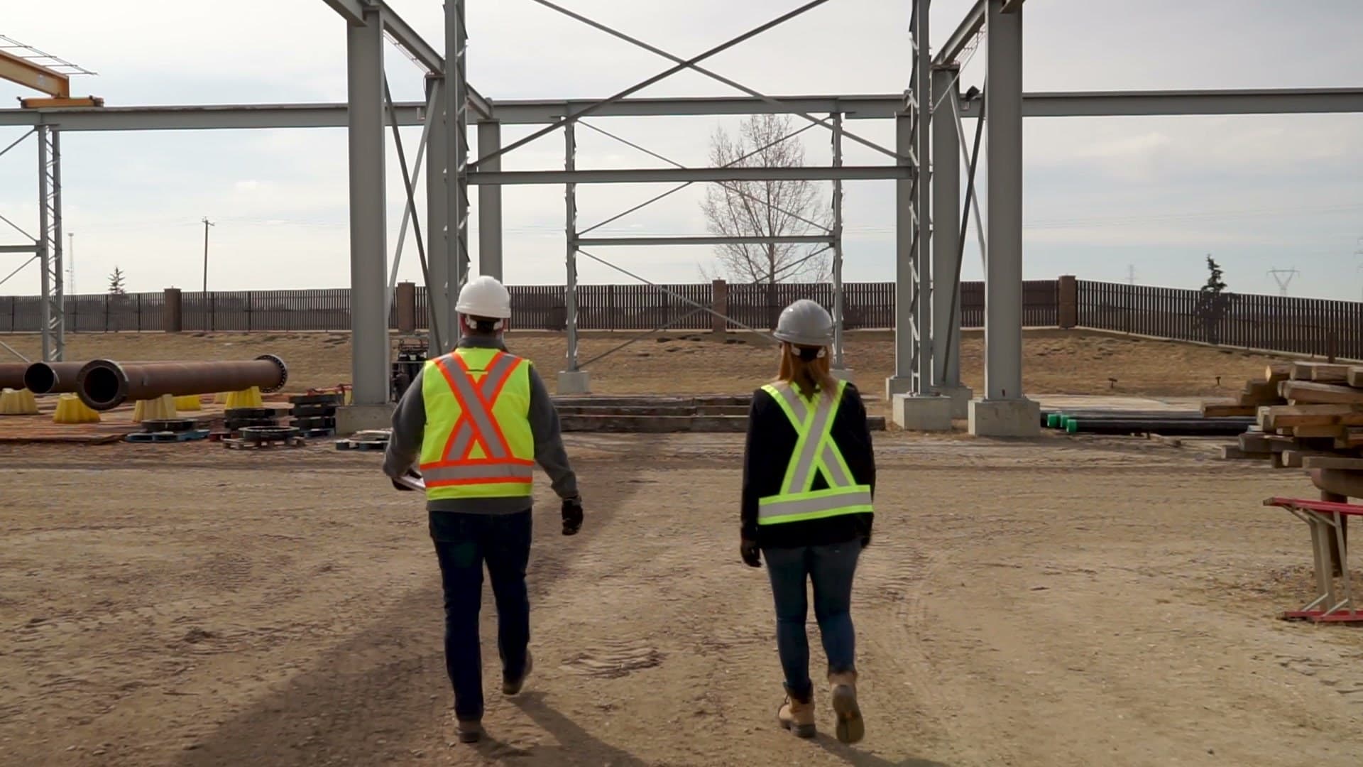 Two safety professionals in hard hats and hi-vis vests walking across a gravel yard between steel building frames.